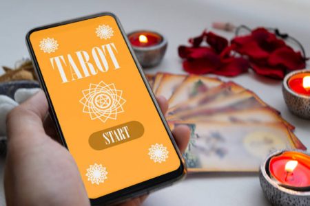 Left hand holding smartphone with tarot app, with cards and different mystical divination objects out of focus. business concept
